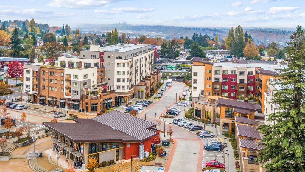 Aerial view of Tukwila, Washington, with modern buildings, parked cars, autumn trees, and Seattle's skyline in the distance under a partly cloudy sky