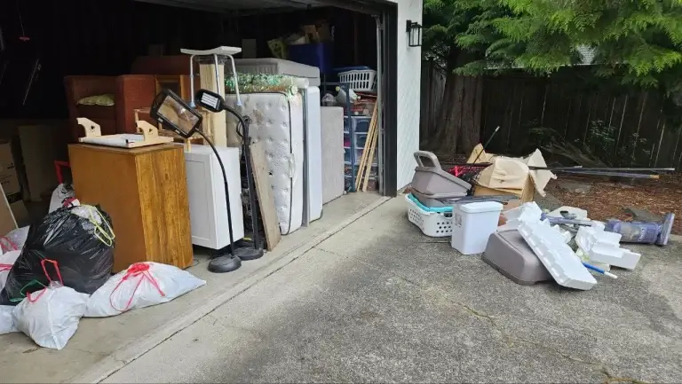 A garage filled with old furniture, mattresses, bags of junk, and containers, creating a cluttered environment.