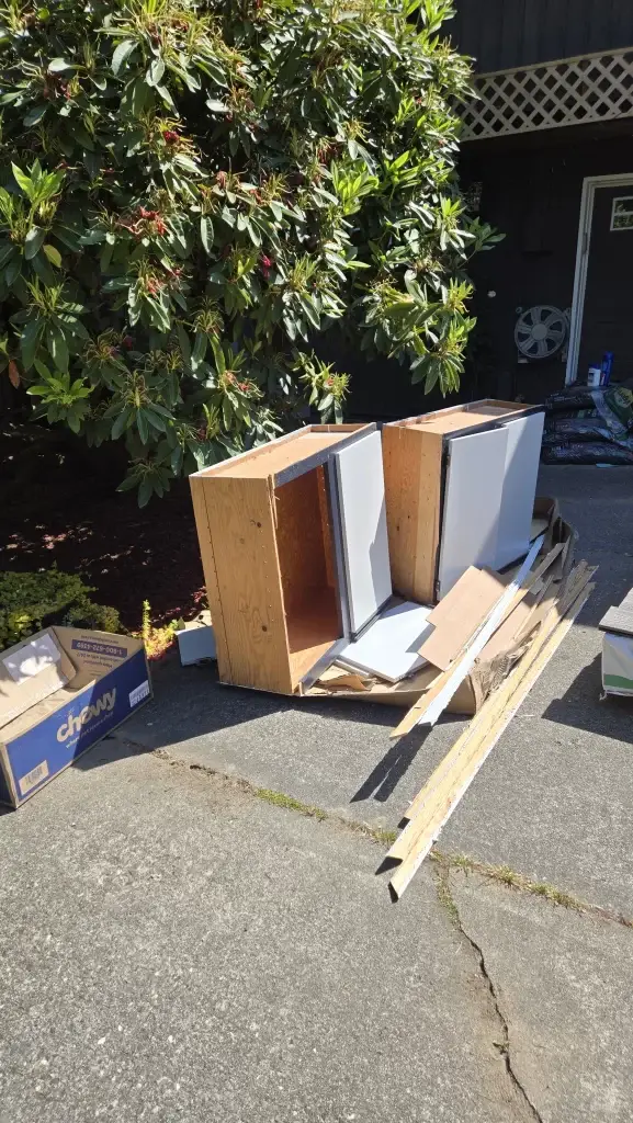 Disassembled kitchen cabinets and debris stacked outside, with a large shrub in the background.