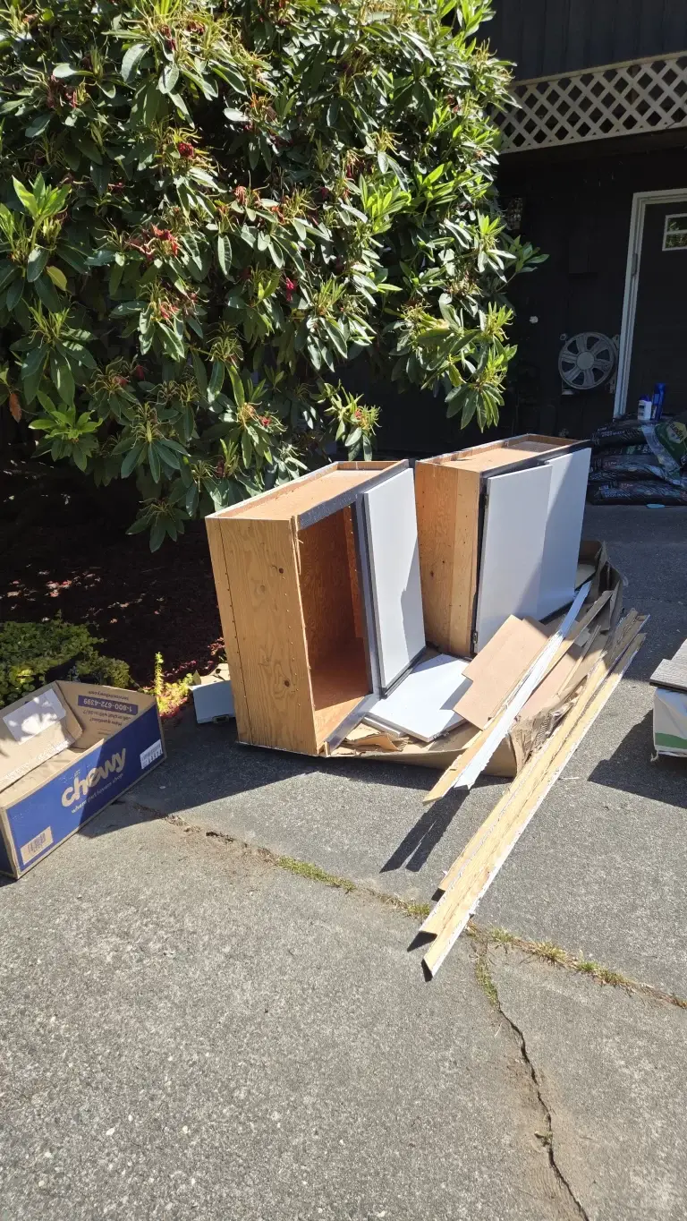 Disassembled kitchen cabinets and debris stacked outside, with a large shrub in the background.