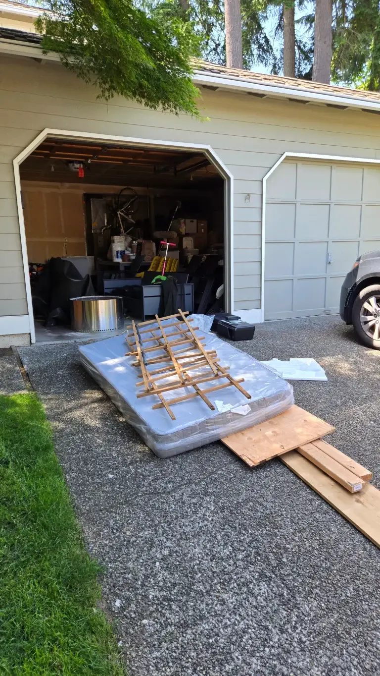 A discarded mattress with a wooden frame and construction debris outside a garage.