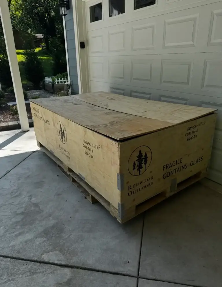 A large wooden crate with fragile glass markings sitting outside a garage.