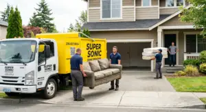 Professional junk removal workers loading a large green couch and a stack of mattresses into a yellow Supersonic Junk Removal truck parked in a residential driveway.